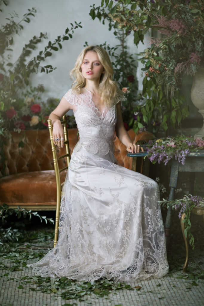 Claire Pettibone wedding dress on blonde model with tossed hair. Sitting on chair with greenery around in natural style.